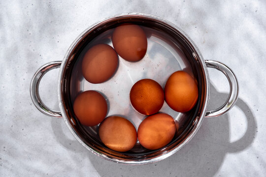 Brown chicken eggs in water inside stainless steel pot, overhead view on light grey concrete background with hard shadows.