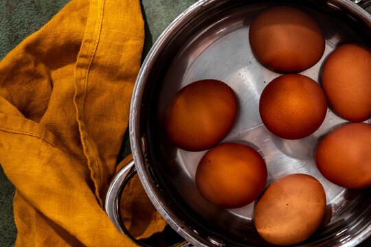Top view of brown chicken eggs submerged in water in pot with ochre napkin, prepared for boiling in bright sunlight.