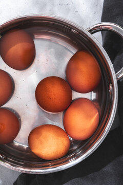 Close up vertical view of brown chicken eggs in water inside stainless steel pot with dark grey towel in hard sunlight.