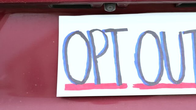 Handmade sign on car license plate slot messages like Opt Out and Data equals money to protest ALPR vehicle surveillance and Fourth Amendment rights