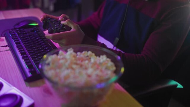 Man holds game controller with hands above keyboard and bowl of popcorn in a studio gaming setup; focused leisure.