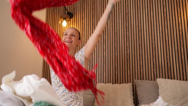 Cheerful young woman sitting on a bed, excitedly throwing colorful clothes into the air while packing her suitcase for a long-awaited summer holiday or travel adventure