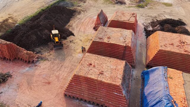 Excavator moving soil beside large stacks of red clay bricks at a brick works