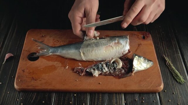 Hands of a person skillfully filleting a fish on a wooden cutting board, showcasing the precise cutting technique and the fish's body positioned with visible entrails and tools nearby