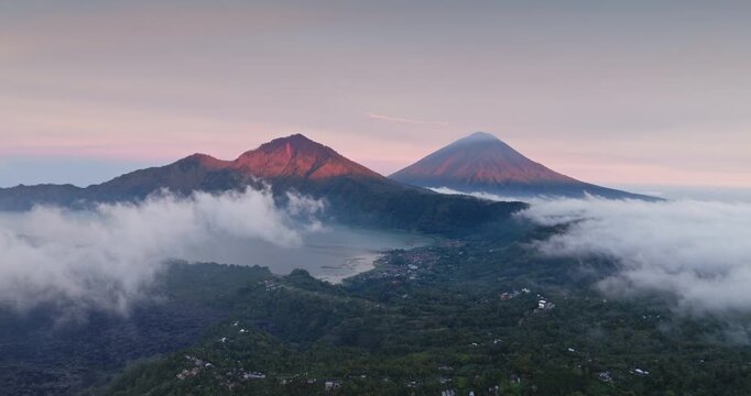 Bali volcanic landscape featuring Mount Batur and Mount Agung volcanoes towering above Lake Batur, surrounded by low morning clouds and lush green forests at a soft sunrise light. Aerial drone flight