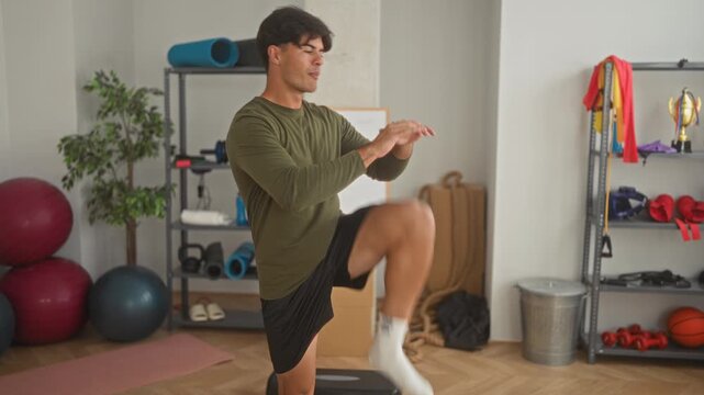 Man performing high knee and leg raise on a step platform with hands on hips in studio near exercise balls, yoga mat and shelving of fitness gear; focus discipline.