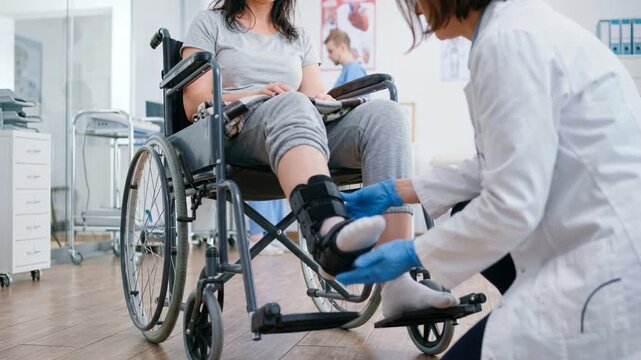 A medical worker assists a client in a wheelchair with leg support. They are at a medical office where the personnel concentrate on rehabilitation. The scene is hectic with other activities