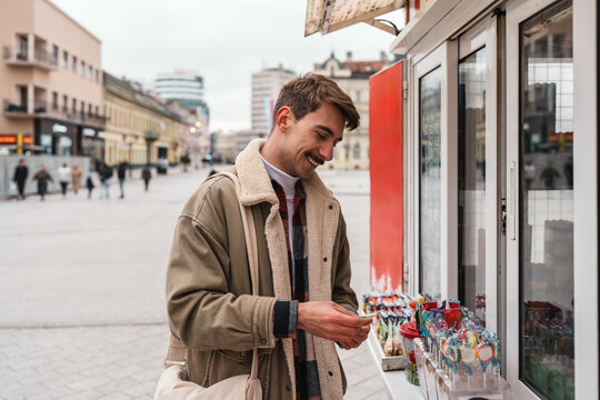A cheerful young tourist holding money while choosing lollipops and candy at an outdoor vendor stand in a European town plaza.