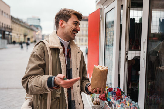 Smiling young tourist holding a bag of warm snacks while interacting with a vendor at an outdoor food stand on a pedestrian boulevard.