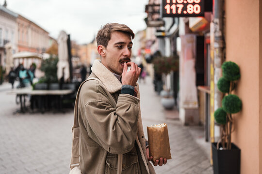 A stylish guy with a mustache enjoys a street food snack during an autumn walk through a European urban pedestrian zone.