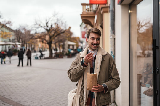 A stylish guy with a mustache holds a paper bag of snacks while strolling along an autumn urban pedestrian avenue.