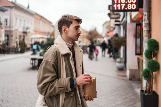 A handsome male tourist wearing a warm shearling coat snacks on popcorn from a paper bag while exploring an old town center past digital shop displays.