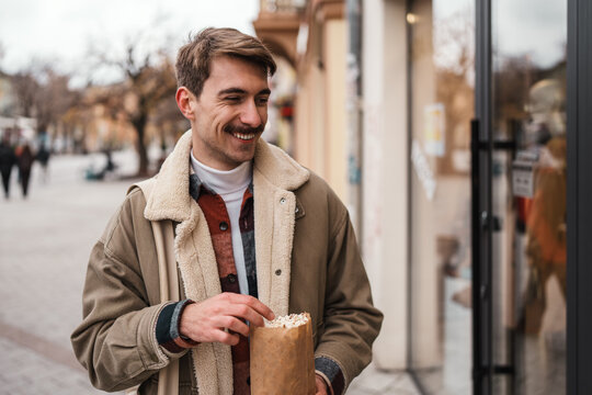 Smiling male pedestrian wearing a warm jacket enjoys a paper bag snack while walking past store windows on an urban sidewalk.