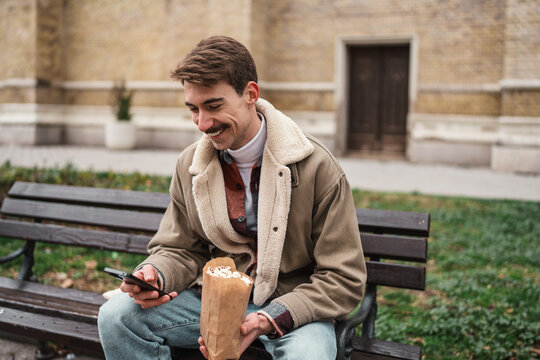 A smiling guy sits outdoors on a wooden city bench, texting on his mobile phone while holding a paper bag of popcorn during an autumn leisure walk.