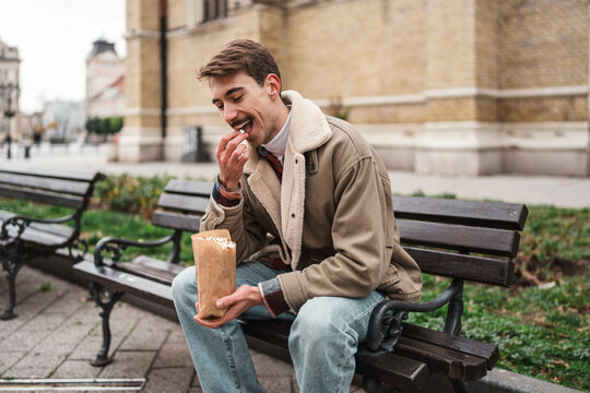 Happy guy enjoying a tasty snack from a paper bag while relaxing on a wooden bench in the city center during a casual stroll.