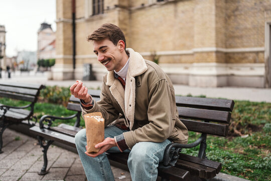 A cheerful guy wearing a warm casual jacket enjoys a crisp paper bag snack during a stroll through a European city square.