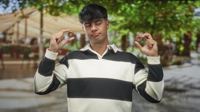 Man in black and white striped shirt holding two biscuits between fingers with a doubtful expression in a street cafe under trees; skepticism playfulness.