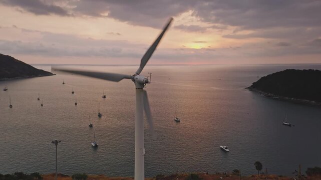 Wind turbine rotating generating renewable energy on a hillside overlooking the ocean with sailboats anchored in a bay during a beautiful sunset on Phuket island, Thailand. Aerial view drone panorama