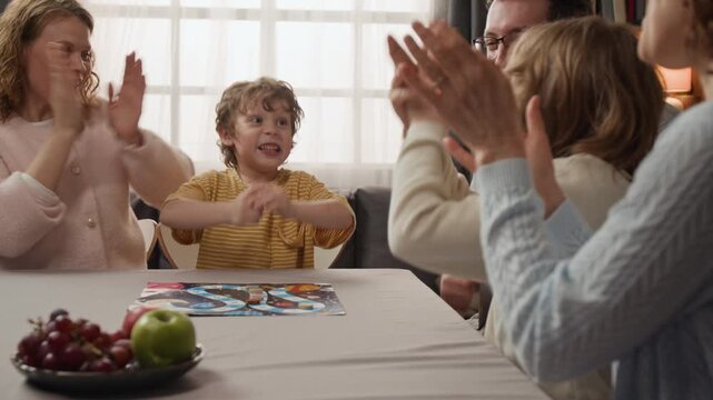 Caucasian family with children and grandmother playing board game at table, young boy celebrating victory while family claps. Ideal for family leisure and home entertainment promos