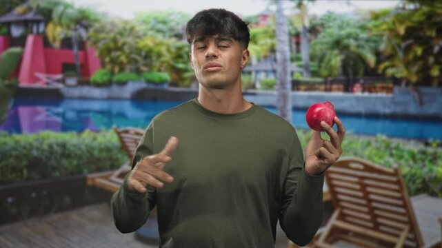 Man holding red apple, grimacing and gesturing with hands on a poolside deck beside a building; uncertainty temptation.