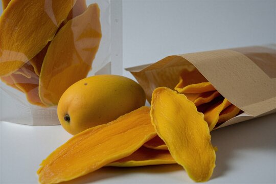 Dried mango slices in craft paper bags on white surface. Fresh yellow whole fruit sits near healthy snack packs. Tropical food pieces ready for eating. Organic vegetarian treats in transparent
