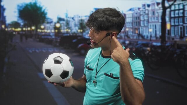 Man referee in turquoise jersey holding a soccer ball and pointing to his headset on a street by canal buildings and lamps; confidence coaching.