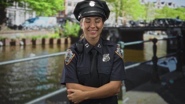 Woman police officer standing with arms crossed and badge visible, smiling while wearing cap by a canal side street and bridge; confidence duty.