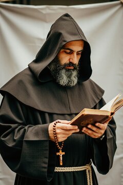 Mature bearded man wears black monk robe with hood. Religious person holds wooden rosary with cross beads reading old holy book. Authentic holy friar studies scripture text during daily spiritual