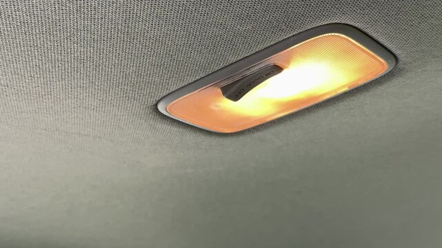  Hand pressing car interior dome light switch on gray ceiling headliner with warm illumination, automotive detail.