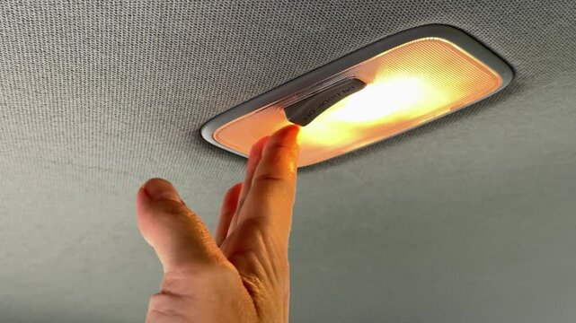 Hand pressing car interior dome light switch on gray ceiling headliner with warm illumination, automotive detail.