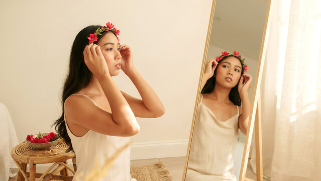 Young woman adjusting floral crown while gazing into full length mirror in bright bedroom interior with reflection wooden mirror headband while looking