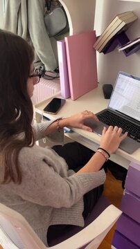 Young female student with glasses studying remotely on her laptop in her room, focused on e-learning and online course work using digital technology for her education