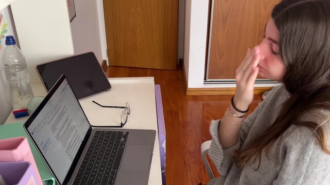 Tired female student yawning and struggling to concentrate while studying online from home showing fatigue and boredom in front of her laptop and tablet in her room
