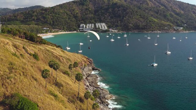 Paraglider flying along the rugged coastline of Phuket, passing over clear turquoise bay filled with yachts anchored near Nai Harn Beach and surrounded by lush green mountains. Summer relax and sport