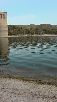Low angle smooth panning video shot of Embalse de Beniarr&eacute;s concrete dam structure reflected in calm water with gentle ripples. Scenic landscape with hills, dry shoreline and yellow buoys in Alicante 
