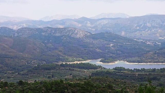 High angle panning video shot of Embalse de Beniarr&eacute;s water reservoir surrounded by green valleys, olive groves and majestic mountain ranges in Alicante province, Spain. Scenic Mediterranean landscape