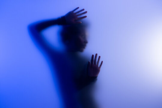 Young black woman posing behind matte glass in studio with blue and lilac light. 
