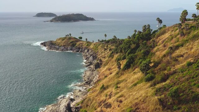 Phuket, Promthep Cape coastal landscape with rocky hills, dramatic cliffs, lush tropical vegetation, palm trees, and expansive turquoise Andaman Sea with a distant island horizon. Aerial drone flight