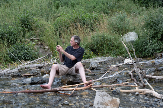 Caucasian man sits on a rock at the base of Stewart Falls and takes a photo with his mobile phone.  Taking photos of a stream in Utah mountains, Mount Timpanogos near Sundance.