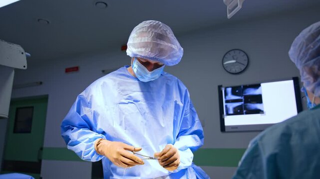 Masked surgeon holding instrument during operation. A clinician in blue sterile attire looks downward under focused lighting.