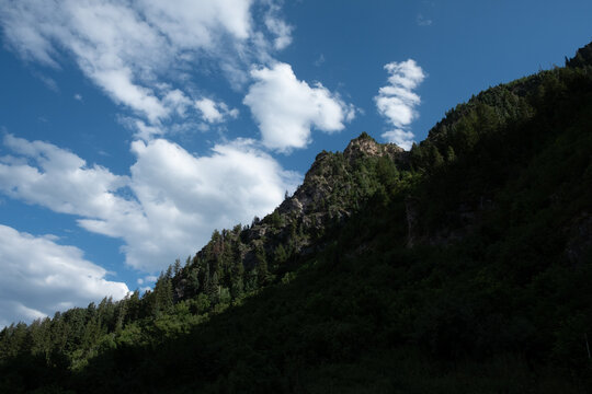 Sunlight on the top of Mt Timpanogos on a summer day with deep blue clouds and fluffy white clouds near Sundance, Utah.  Tall rocky mountain covered with pine trees half covered in the shadows.