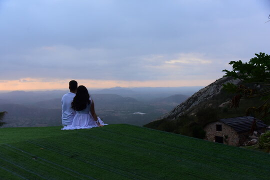 romantic couple sitting on hilltop enjoying mountain vista view at sunset, man and woman dressed in white sitting on green grass looking at horizon, lovers embracing at scenic mountain viewpoint dusk 