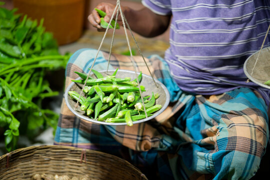 A street vendor holding a traditional balance scale with fresh okra in a market
