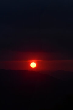 dramatic red sun setting over mountain horizon, glowing solar orb against dark sky at dusk, minimalist nature landscape sunset concept, vibrant sun disk appearing between dark clouds and hills