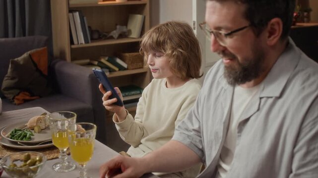 Caucasian child using smartphone during family dinner and talking to Caucasian father. Suitable for advertising on screen time limits and modern parenting