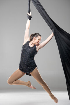 Aerial silk acrobat performing dynamic split pose, woman in sparkling black leotard suspended in black fabric, smiling expression, studio shot on gray background