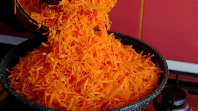 Adding a large portion of grated carrots into frying pan for cooking, preparing fresh vegetables for soups, stews and homemade meals, healthy food preparation in kitchen