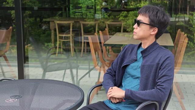 Man sits calmly outdoors, wearing sunglasses and a navy jacket. He rests hands together, gazing thoughtfully beyond the glass. Setting suggests quiet contemplation in a garden cafe