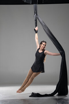 Aerial silk acrobat performing dynamic split pose, woman in sparkling black leotard suspended in black fabric, smiling expression, studio shot on gray background