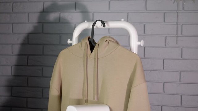 Woman steams clothes. Person steaming a beige hoodie with a portable iron steamer against a gray brick wall, performing household chores